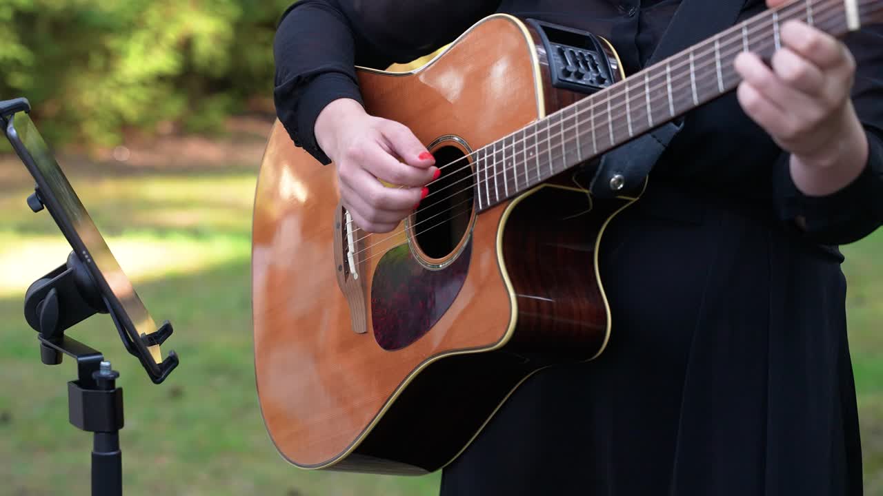 Close-up of a woman playing an acoustic guitar outdoors, dressed in black, with a music stand and greenery in the background—perfect for live music or performance-related themes
