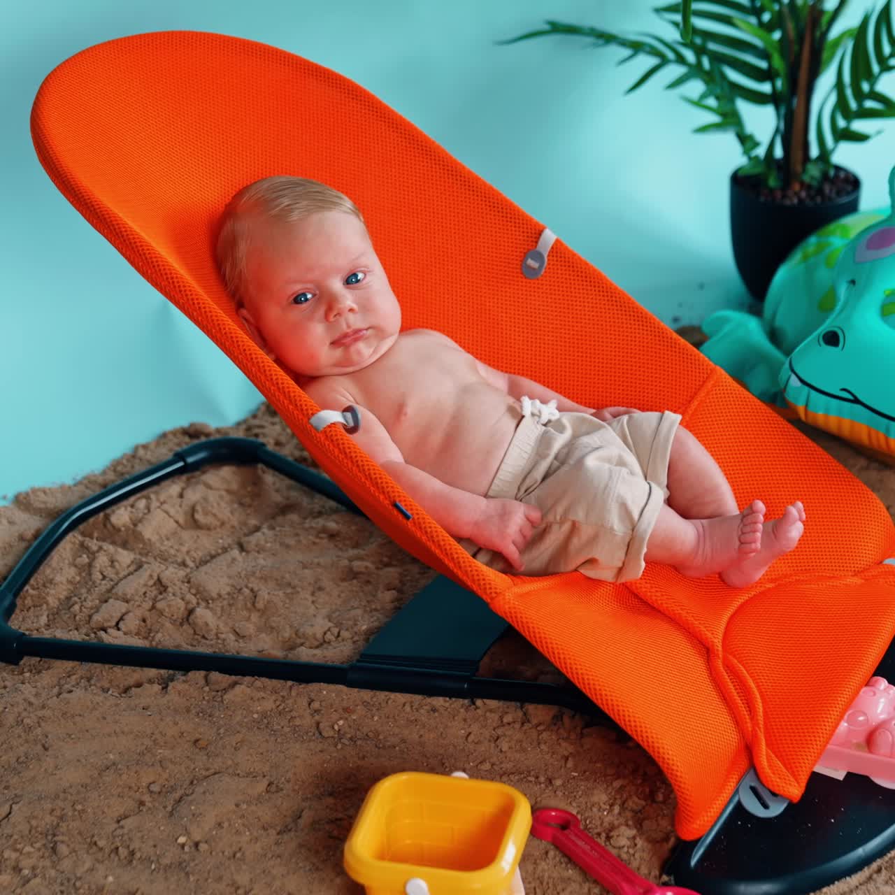 Blond infant boy lies in the orange chair relaxing. Happy vacation on the imitated beach. Side view