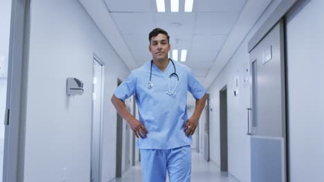 Portrait-of-smiling-mixed-race-male-doctor-wearing-scrubs-walking-in-hospital-corridor