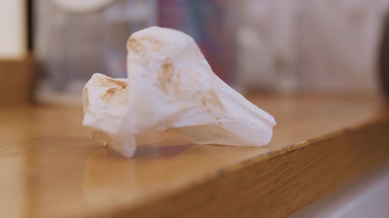 Fazzoletto di carta accartocciato su un piano di lavoro in legno macchiato, tovagliolo usato con briciole e macchie di unto, primo piano con profondità di campo ridotta che mostra venature e texture, il dopo-mattina in cucina, illuminazione naturale, pronto