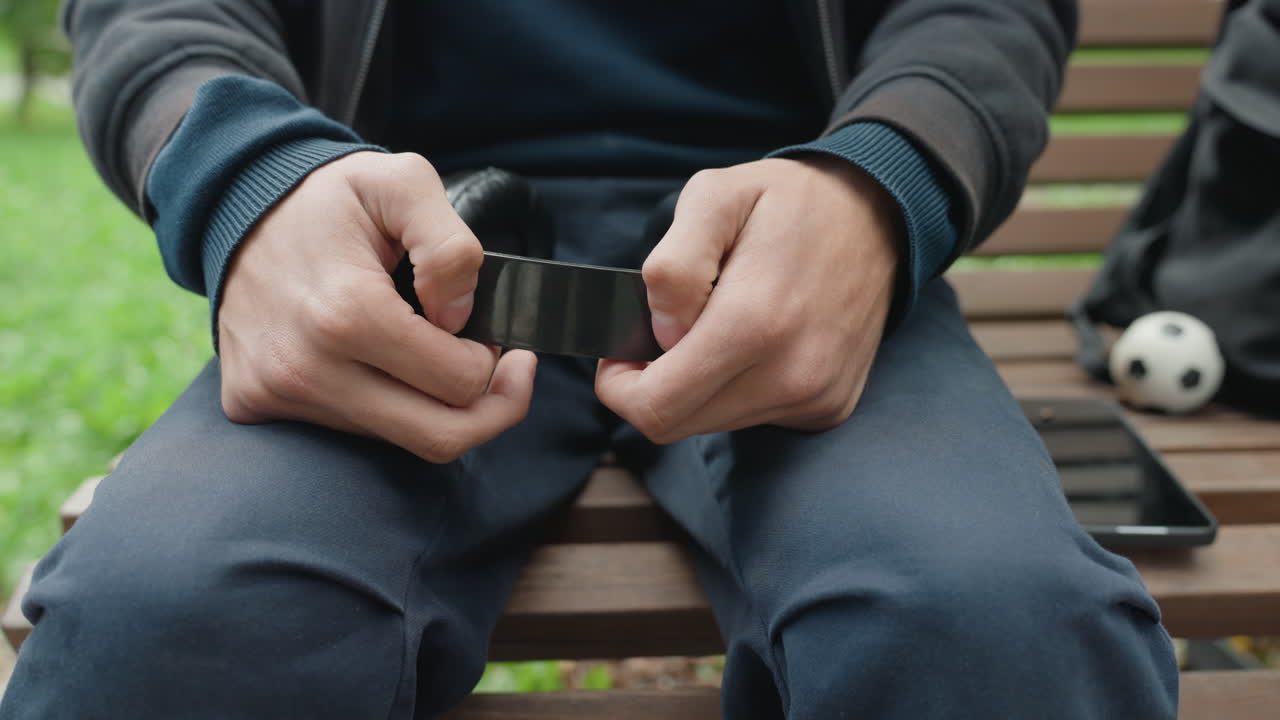 uomo seduto su una panchina che tiene una manetta, primo piano delle mani che slacciano la costrizione nera, zaino e pallone da calcio accanto al sedile, tablet sulla panchina, atmosfera esterna nuvolosa con tono teso e pieno di speranza