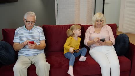 abuelos y nieta sentados en un sofá rojo juntos y mirando sus teléfonos