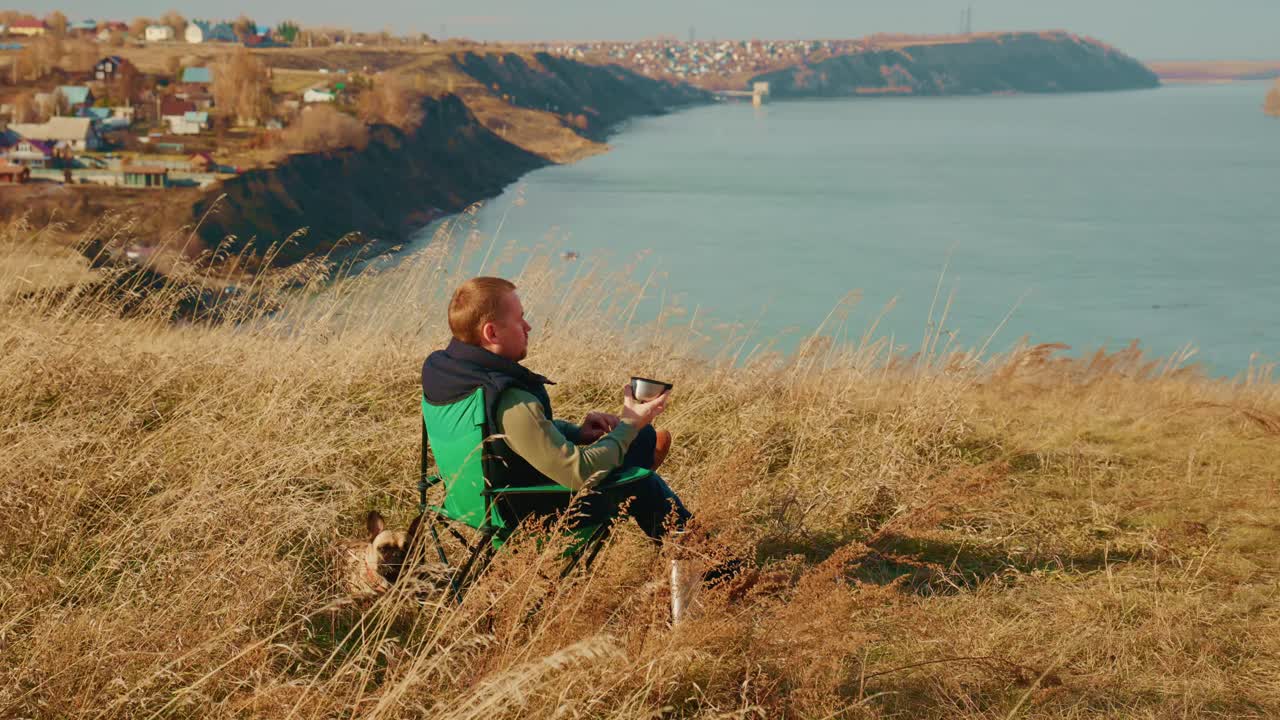 A contemplative individual seated on a grassy hillside overlooking a serene river, taking in the picturesque landscape with a backdrop of quaint townhouses dotting the distant shore, reflecting on nature's tranquility