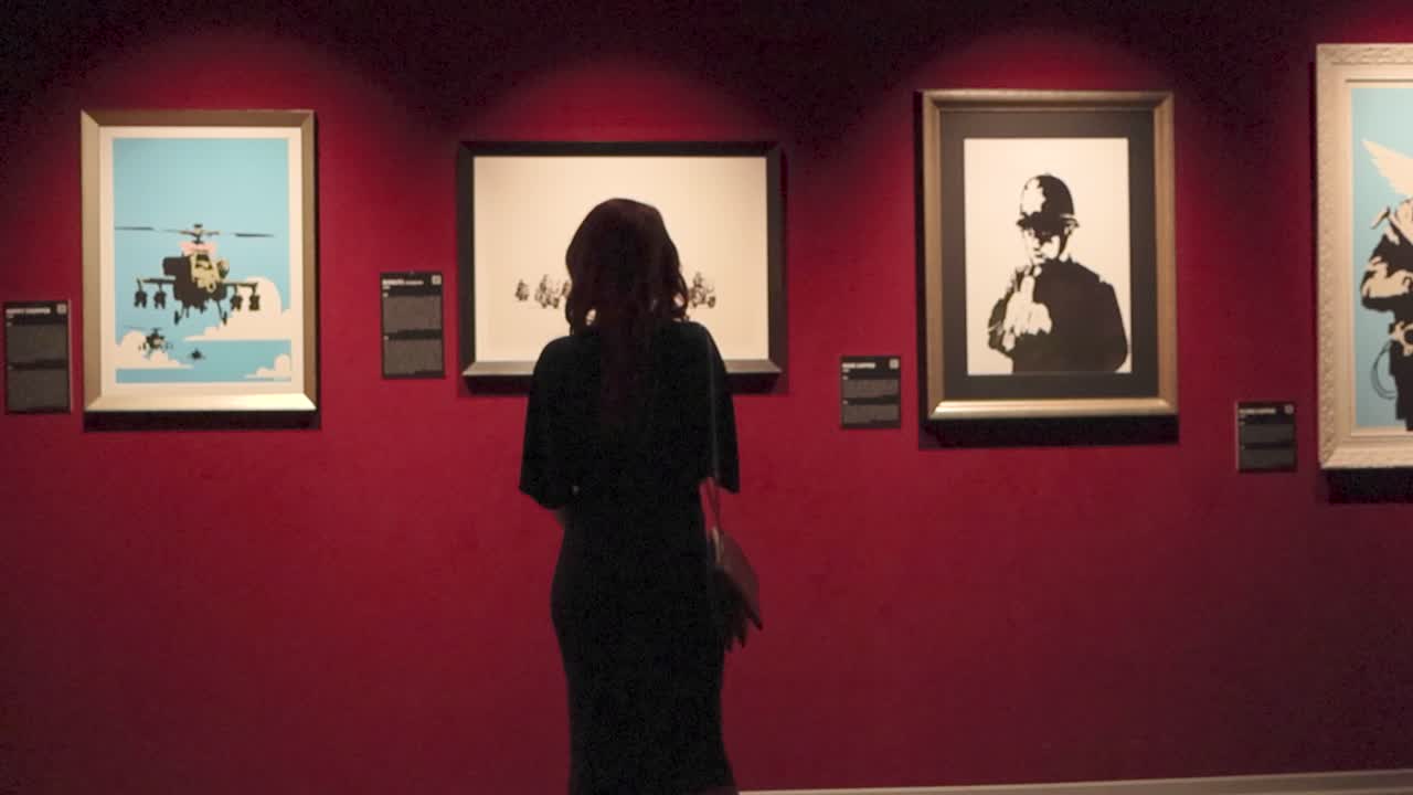 Gorgeous woman at a Banksy exhibition watchin multi-layered stencil art made by him. The woman has curly hair and a dress while carrying a purse with her indoors. Art on red wall that is well lit.
