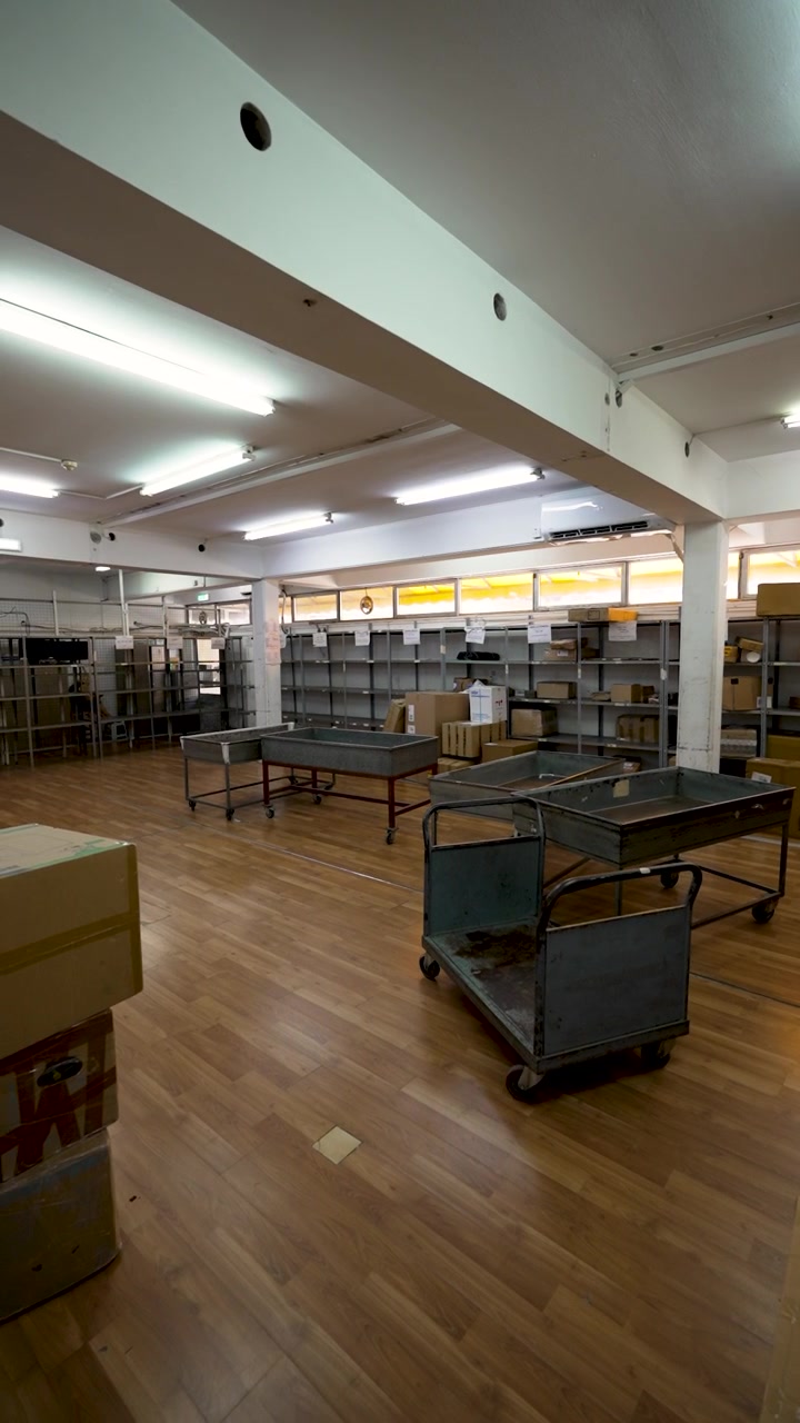Boxes are arranged on shelves in a warehouse. Carts are placed in the middle of the room. Lights are on, creating a bright space for organizing goods.