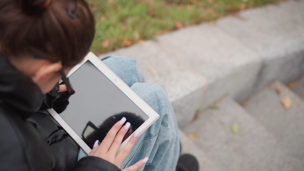 Primo piano di giovane donna con occhiali e abbigliamento casual che tiene un tablet elettronico in grembo mentre è seduta all'aperto. Le sue mani con unghie lunghe interagiscono costantemente con il dispositivo.
