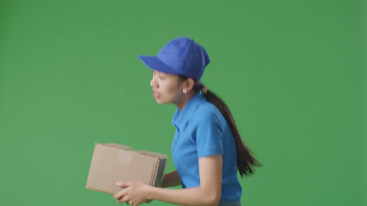 Side View Of Asian Female Courier In Blue Uniform Running While Delivering A Carton On Green Screen Background In The Studio
