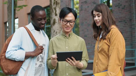 camera zoom out on caucasian woman, asian and african american students talking while looking at the tablet in the street near the college