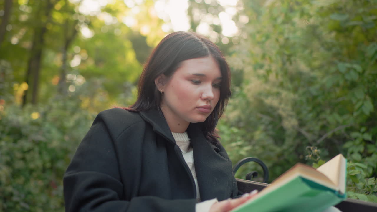 joven concentrada sentada en un banco del parque con las piernas elevadas, leyendo un libro verde y ajustándose el pelo entre las páginas, con un vaso de papel en el suelo, luz suave de otoño, ambiente tranquilo de estudio al aire libre con un abrigo cómodo y pantalones blancos