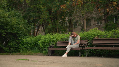 young woman sits alone on wooden bench in tranquil park, resting head on arm in reflective, somber posture. she s surrounded by lush greenery and trees