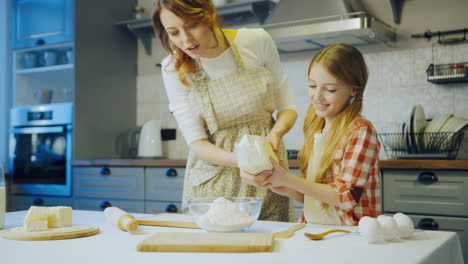 beautiful young mother preparing everything for cooking, pouring the flour in the bowl and her daughter helping her. nice modern kitchen. indoor