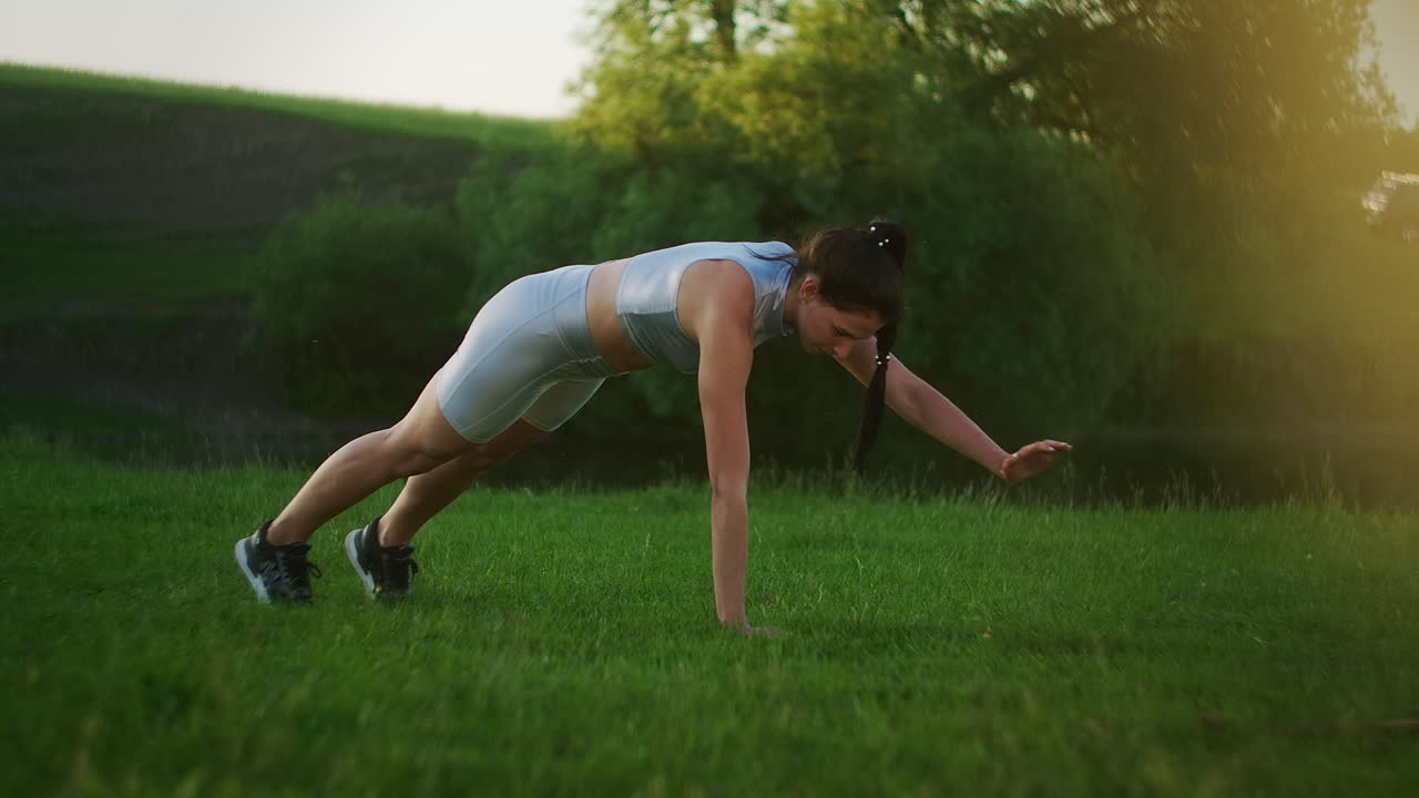 a woman in a Park on the grass stands in the plank exercise and raises her right and left hand in turn. static exercise on abs