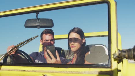 happy caucasian couple sitting in beach buggy by the sea using smartphone