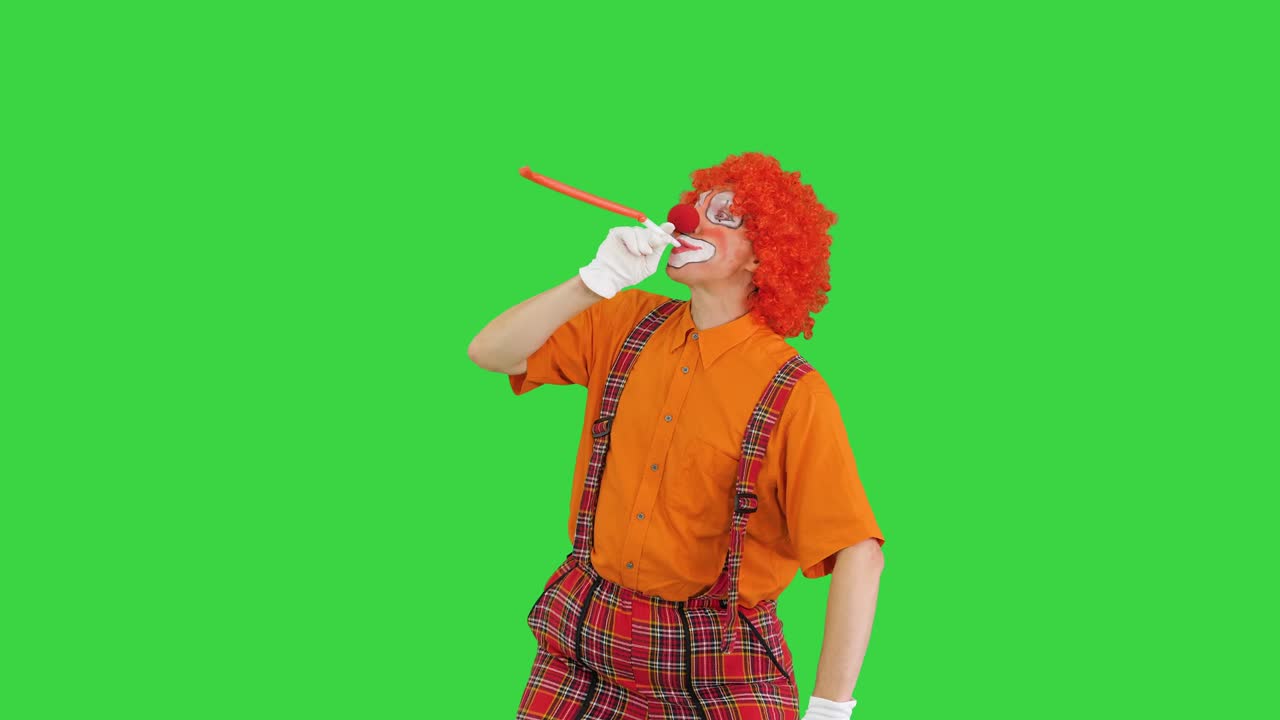 Clown con naso rosso che soffia in una trombetta da festa su green screen, chroma key.