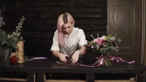 the woman florist completes the composition of the bouquet, cuts off the tips of silk ribbons with scissors. excited of her work. front view. wodden interior background