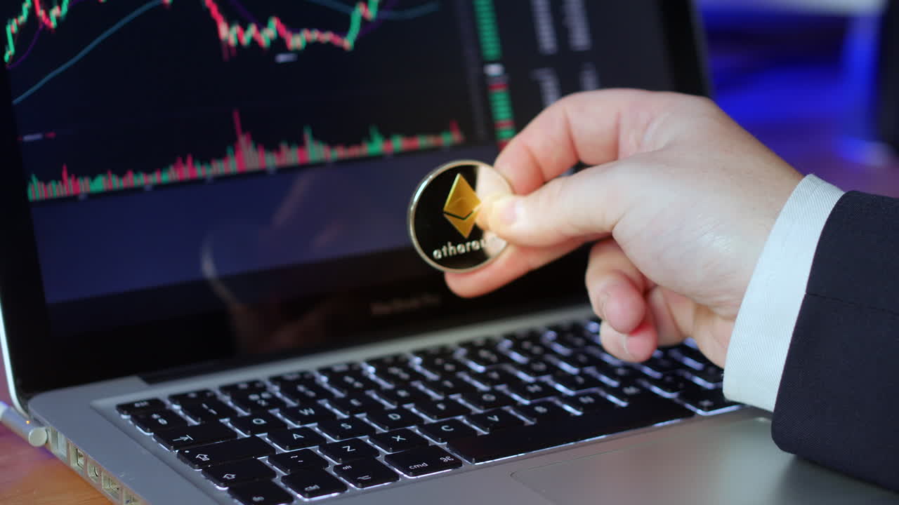 l'uomo d'affari capovolge la moneta ethereum tra le dita mentre guarda i grafici di scambio e i prezzi su un laptop
