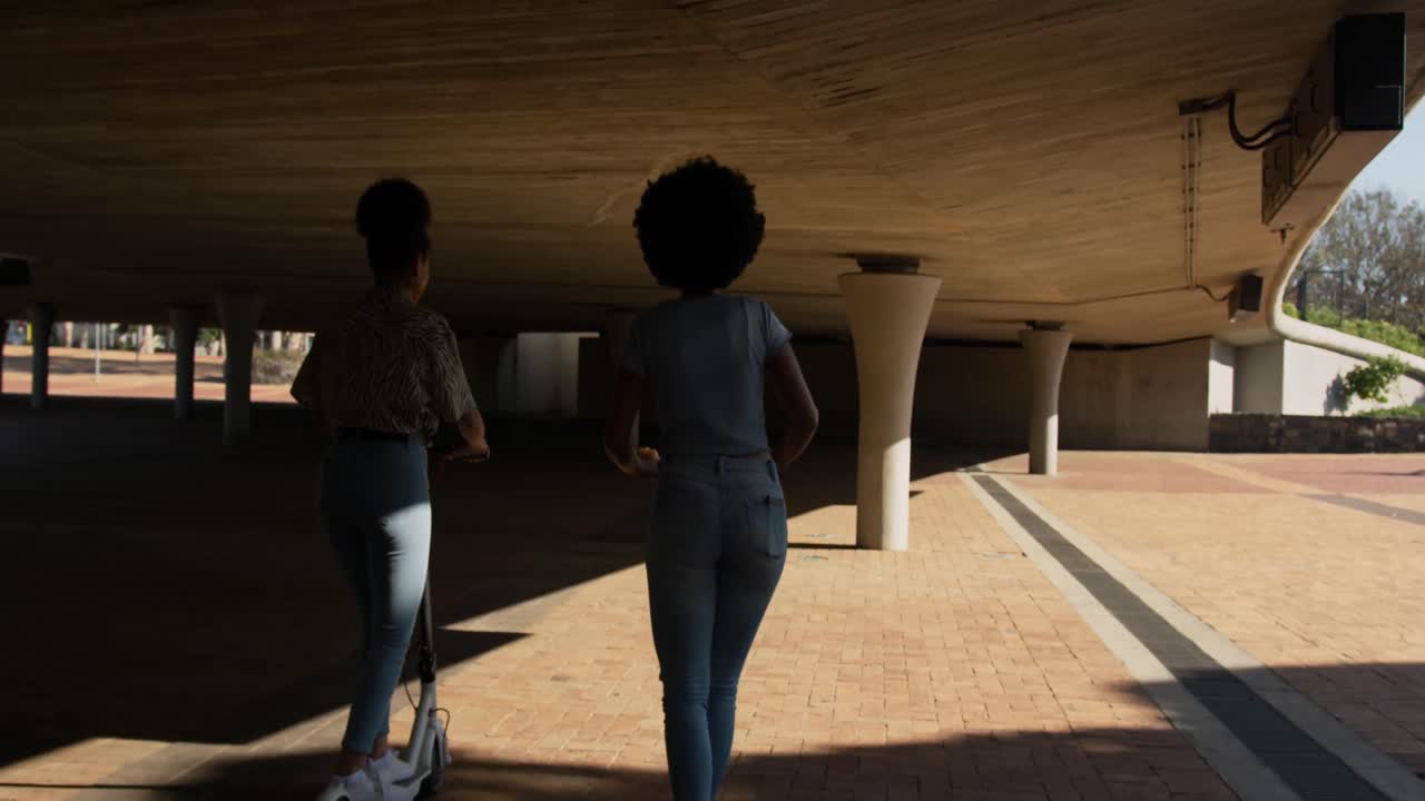 Two mixed race women riding electric scooter under bridge