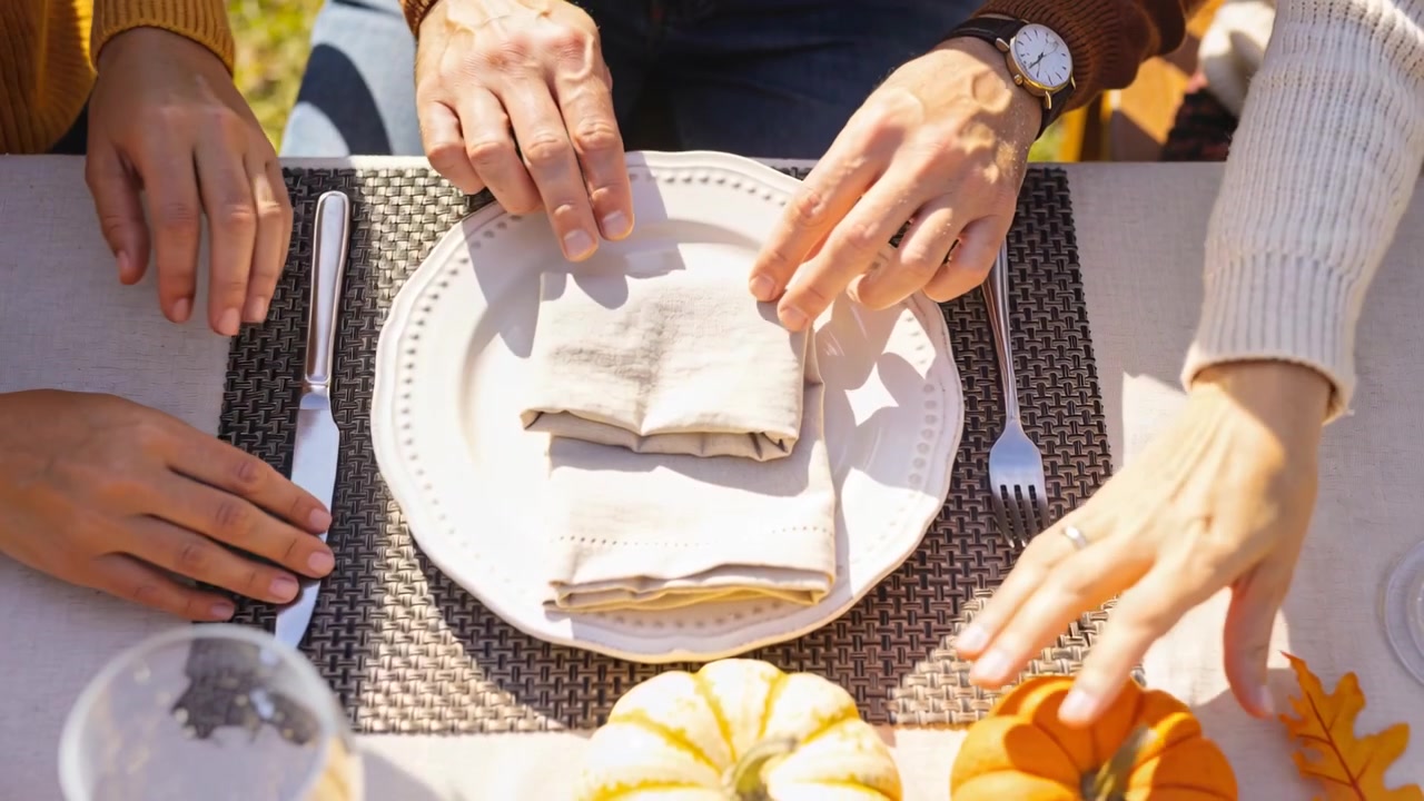Lifting yellow-knit hand reshaping napkin at patio, others aligning fork, placing pumpkin for meal