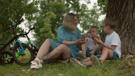 a mother sits on the ground under a tree, offering a snack to her two children, both kids eagerly accept the treat with happiness, sitting on a blanket in the park