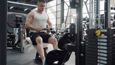fitness trainer seated on bench pulls cable row handle with focused effort in busy modern gym environment draped towel on bench weight stack visible showing dedication to strength and muscle growth