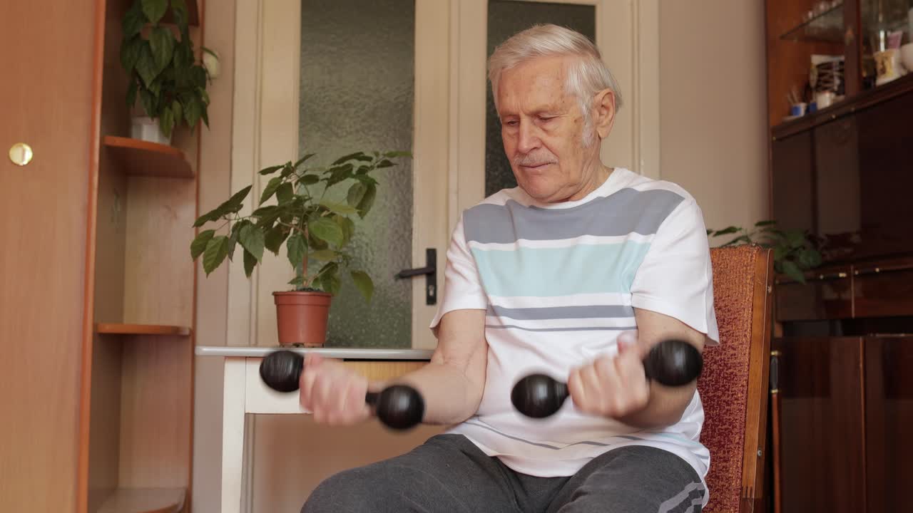 고령 백인 남자가 집에서 몸무게를 들어올리는 덤벨 운동을 하고 있습니다.