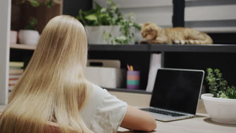 a teenage girl writes at a desk with a laptop. her cat is sitting on the windowsill nearby.