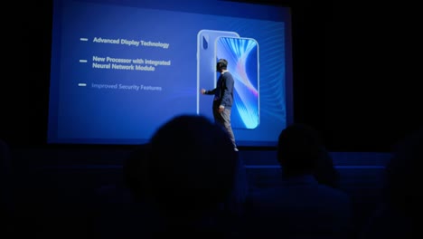 live event with brand new products launch: keynote speaker reveals smartphone device to audience. movie theater screen shows mock-up touch screen mobile phone with high-end features and top highlights