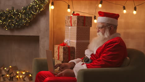 Der-Weihnachtsmann-Arbeitet-An-Einem-Laptop-Und-Sitzt-Zu-Hause-Auf-Dem-Sofa-Vor-Dem-Hintergrund-Von-Weihnachten.-Der-Weihnachtsmann-Antwortet-Auf-Die-E-Mails-Der-Kinder.-Hochwertiges-4K-Filmmaterial