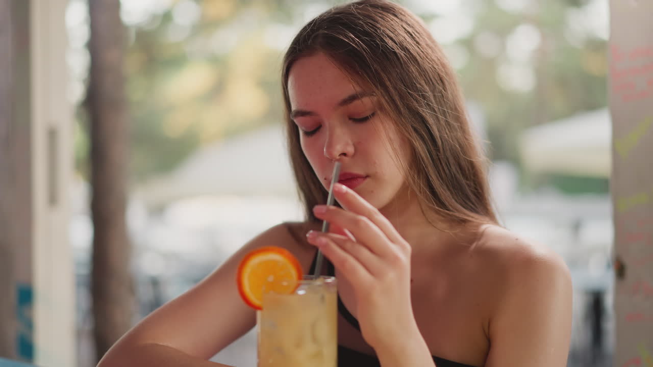 Tired long-haired woman stirs cocktail with straw in bar. Exhausted lady drinks alcoholic beverage after hard working week in beach bar. Weekend relaxation