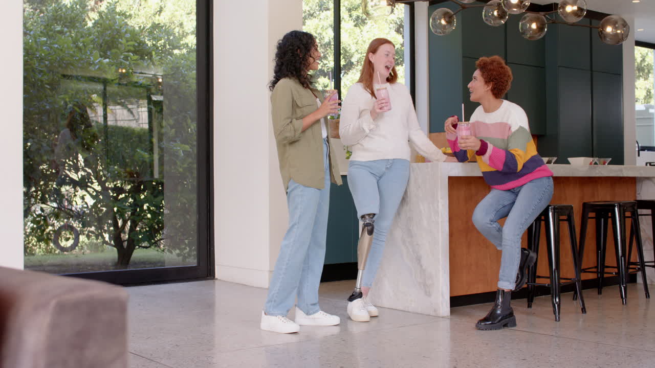 Chatting and laughing, diverse female friends enjoying drinks together in modern kitchen, copy space