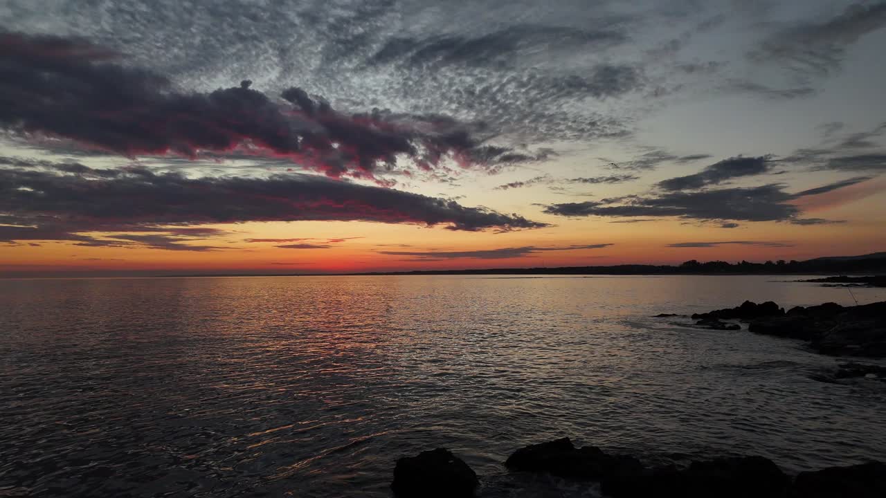 In Maldonado, Uruguay, just after sunset, the calm sea sends gentle waves between coastal rocks. The sky still holds warm tones, and the water reflects the fading light of day