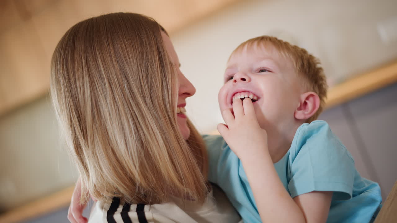 Una madre guarda un ragazzino seduto sulle sue ginocchia mentre il bambino la guarda con un'espressione luminosa; la donna lo tiene delicatamente, tenero momento di cura, calore e amore in un'accogliente cucina moderna
