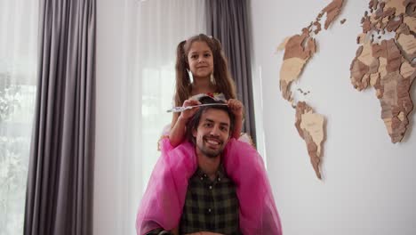 portrait of a happy family little brunette girl in a pink fairy costume with a magic wand sitting on the shoulders of her happy brunette male father in a green checkered shirt in a modern home