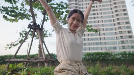 young pretty asian woman showing happy life in city, positive energy, happy joyful girl expressing overwhelming happiness, looking at camera sitting against modern high rise condo green garden park