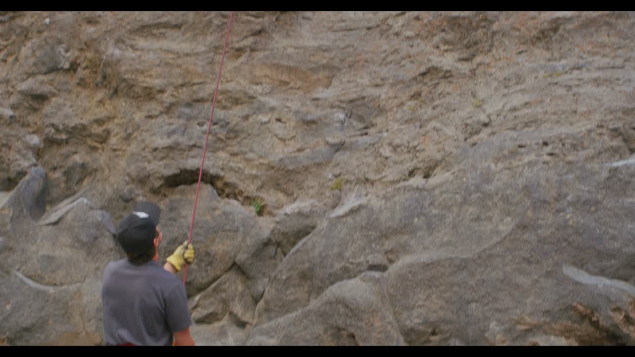 The camera tilts up from a climbing partner on the ground to reveal a rock climber high on a rock wall