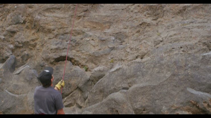 The camera tilts up from a climbing partner on the ground to reveal a rock climber high on a rock wall