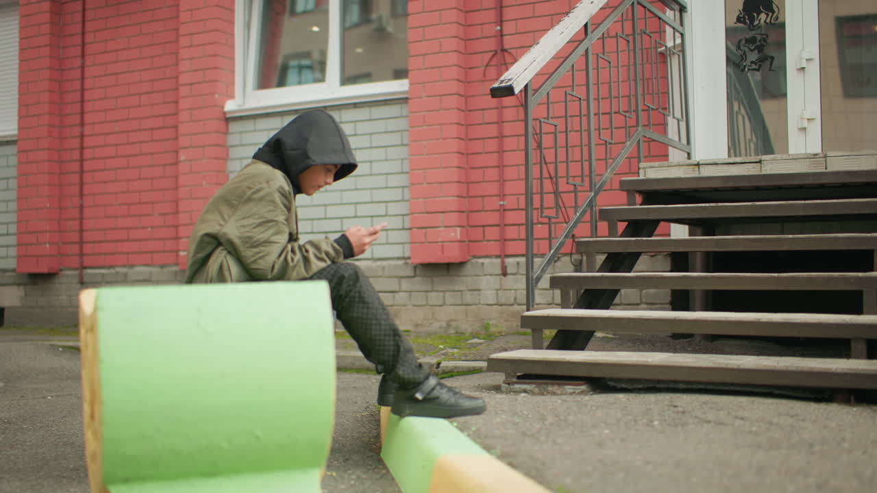 Side view of young boy in hooded jacket seated outdoors on green bench placing leg on wedge, focused intently on handheld device, with red brick building