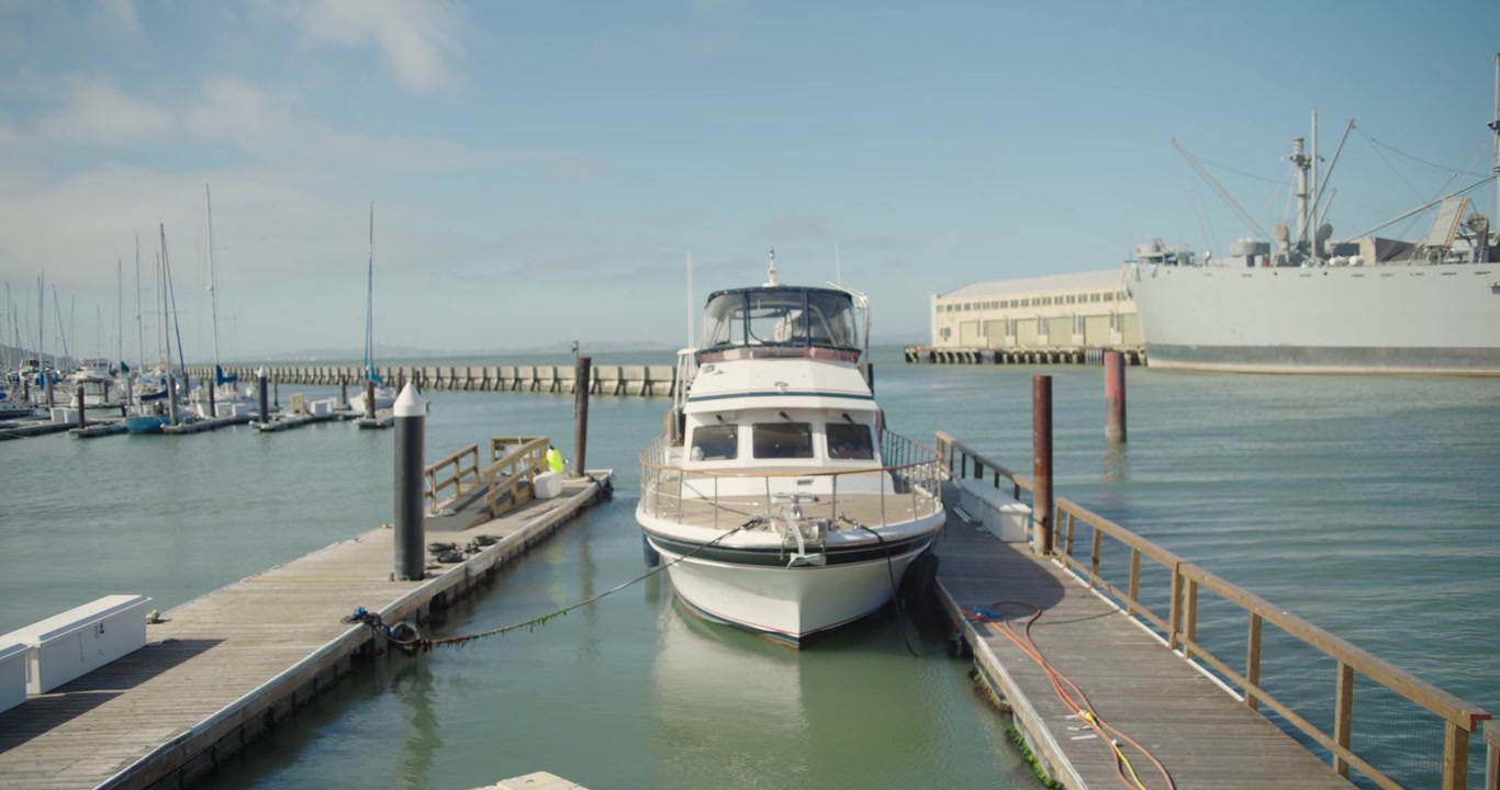 Ripresa cinematografica 4K di un piccolo yacht ormeggiato a un molo di San Francisco. Barche a vela e una nave navale storica creano una tranquilla scena portuale sul lungomare con acque calme della baia