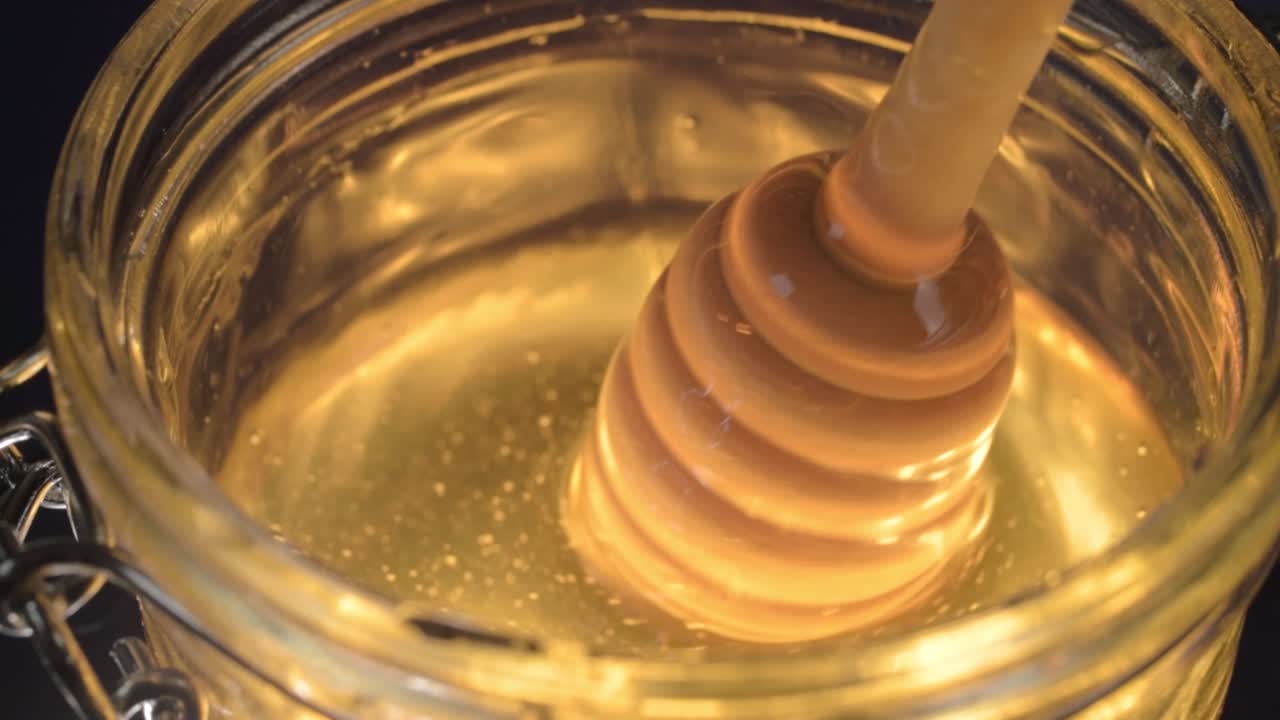 Stirring golden runny honey in a glass jar with honey drizzle spoon