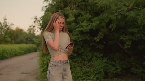 mujer con la parte superior recortada revelando su vientre, caminando por un camino rodeado de exuberante vegetación mientras usa su teléfono, ajusta su cabello con la mano derecha, centrado en el dispositivo