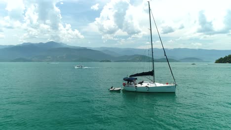 Aerial-drone-orbiting-shot-of-moored-sail-boat-in-a-turquoise-calm-ocean-with-people-inside-the-boat-getting-it-ready-to-sail-in-a-beautiful-tropical-horizon-with-mountains