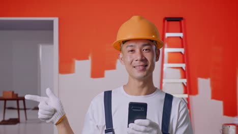 close up de un pintor asiático que usa un casco de seguridad usando un teléfono inteligente y apuntando a un lado mientras está de pie con la pared de pintura en la habitación