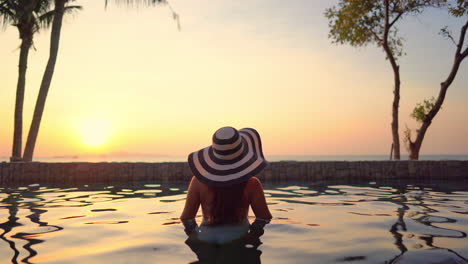 Female-tourist-wearing-a-floppy-hat,-standing-in-the-swimming-pool-being-half-submerged-in-the-water-and-admiring-the-sunset