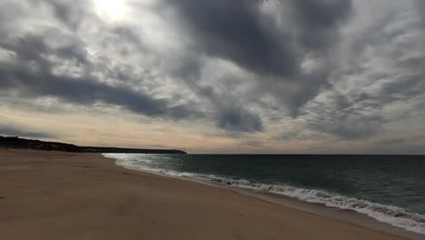 Dramatic-wide-shot-of-golden-beach,-ocean-shore-and-hiding-sun-behind-dark-cloudscape-at-sky