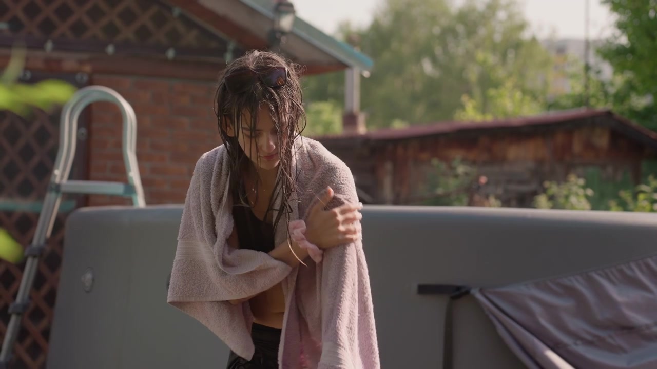 Young woman with wet hair dries off after swimming wrapping pink towel around herself in outdoor backyard setting. Woman wears black swimwear and sunglasses on her head while standing