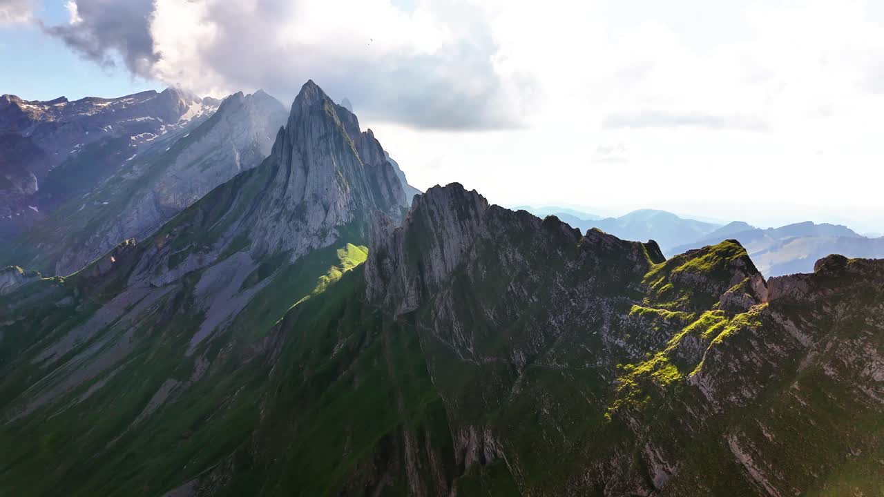 Aerial view of Schäfler Switzerland mountain ridges, highlighting dramatic rocky peaks, steep slopes, lush greenery, and alpine sunlight breaking through clouds over the jagged summit formations