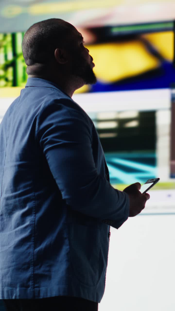 Uomo afroamericano che usa lo smartphone davanti a grandi schermi digitali.