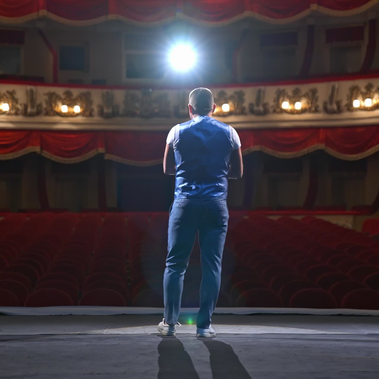 Performer speaks in front of beautiful richly decorated theater hall. Rows of red chairs in theater hall. Big empty theater with one person on stage