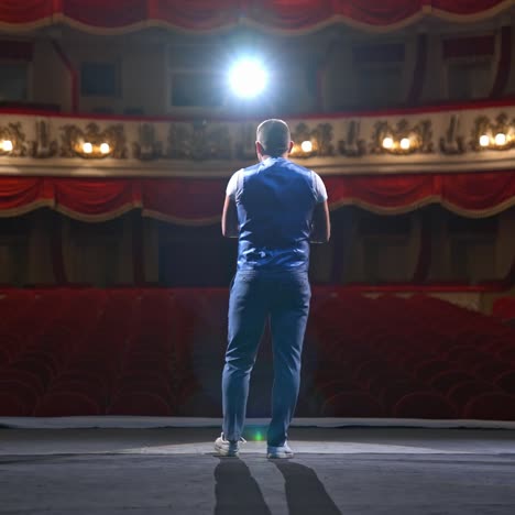 performer speaks in front of beautiful richly decorated theater hall. rows of red chairs in theater hall. big empty theater with one person on stage