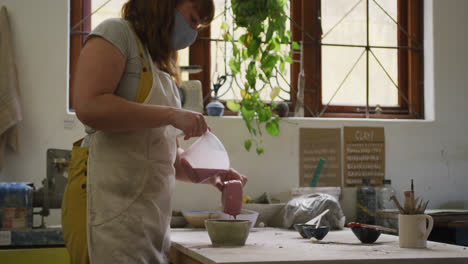 female caucasian potter wearing face mask pouring paint on pot at pottery studio